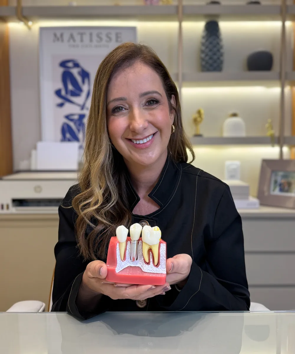 Dra. Micheli Bragantini sorrindo no consultório da Oral Concept, com um modelo de implante dentário nas mãos